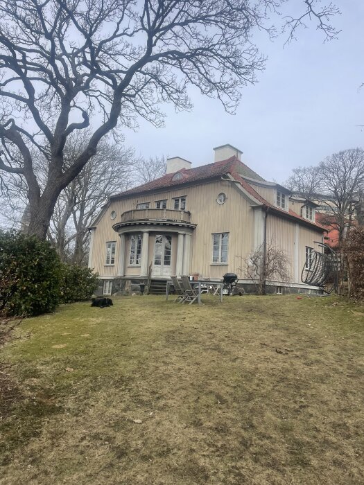 Stort trähus med rött tak i stadsmiljö, omgiven av träd och ekar. Några utemöbler och en grill står på gräsmattan framför huset.