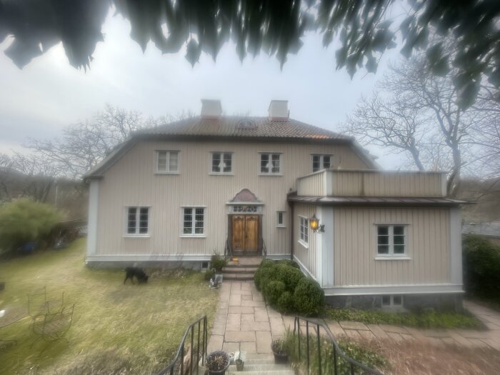 Stor villa i stadsmiljö med beige fasad och tegeltak, omgiven av grön tomt och stora träd. Hunden syns på gräsmattan.