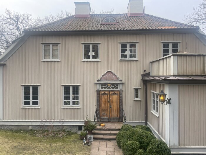 Stort trähus i beigt med dubbeldörrar i trä, omgiven av buskar och ekar. Finns i stadsmiljö, fasaden delvis skuggad.