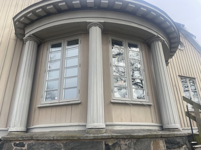 Bågformad fasad på hus med stora fönster och grå pelare, liggande i stadsmiljö med träd reflekterade i glaset.