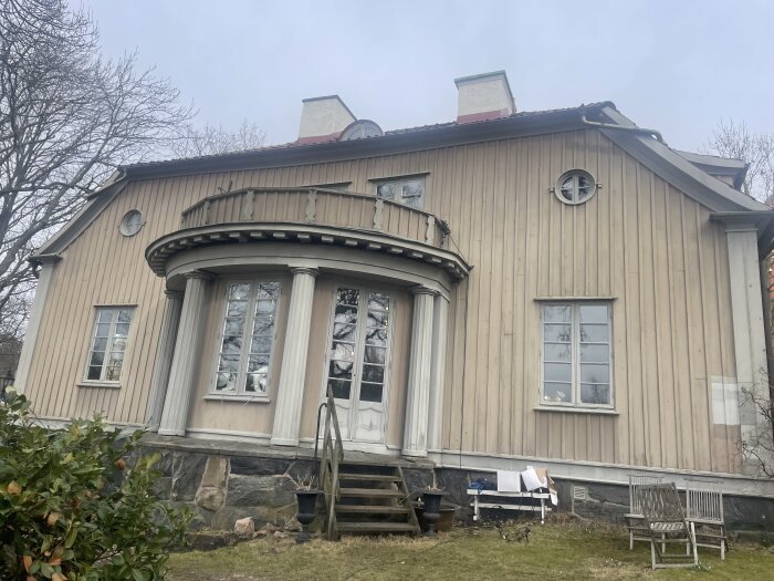Trähus med klassisk design, flera fönster och trappa, omgiven av träd och gräsmatta. Fasaden är beige och huset har runda och fyrkantiga fönster.