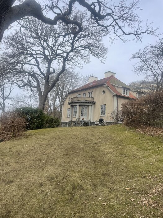 Stort hus med beige fasad och rött tak, beläget på en gräsmatta omgiven av nakna trädgrenar, mitt i en stadsmiljö.