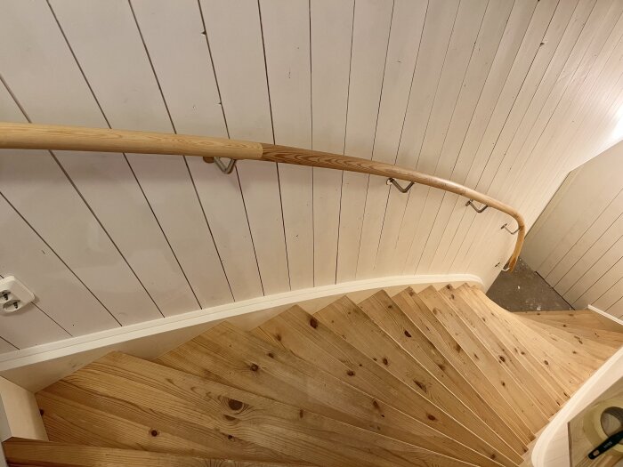 En trapp med en trehåndløper festet til veggen med metallbeslag. Veggen er panelkledd, og trappen består av tregulv i lys farge.