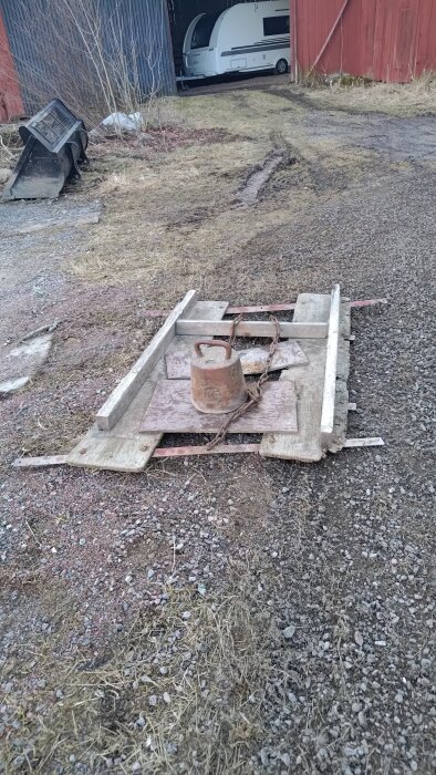 Slitet vinkeljärn på marken, omgiven av träplankor och en kedja med en metallvikt ovanpå, på en grusig gårdsplan.