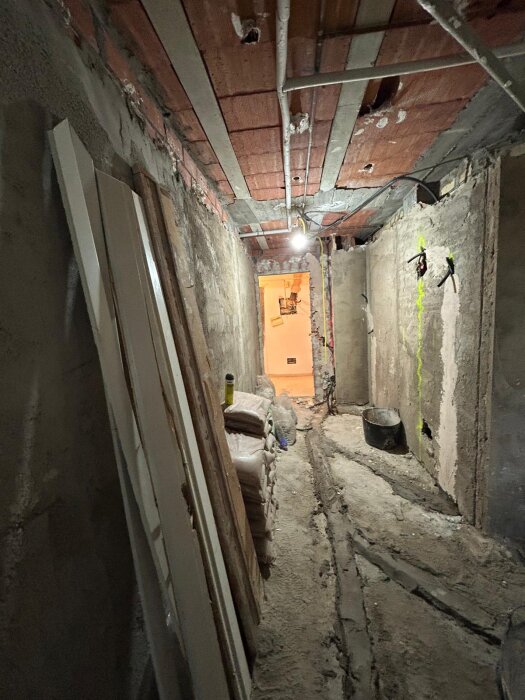 Renoveringsprojekt med en lång, ofärdig korridor. Taket har synliga rör och golvet är ojämnt, med staplade byggmaterial längs väggarna.