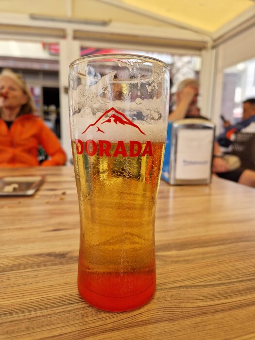 Ett glas öl med märket Dorada på ett kafébord, personer i bakgrunden i San Sebastians solsken.