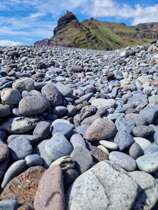 Klippor och ett berg i bakgrunden med en strand täckt av rundade, stenar i förgrunden under en blå himmel med moln.