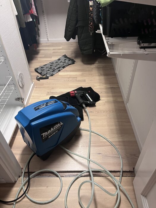 En garderob med trägolv, Elfa hyllor och en blå Makita kompressor. Kläder hänger på rad, en mattbit på golvet.