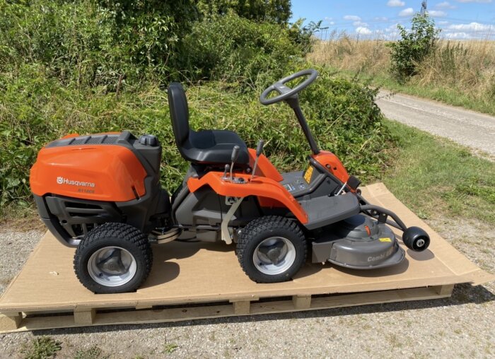Husqvarna åkgräsklippare på pall vid en grusväg, omgiven av grönska och under klarblå himmel.