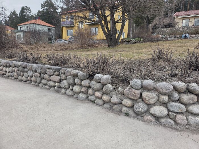 Stenmur vid villaträdgård med buskar ovanpå, gräsmatta och trähus i bakgrunden.
