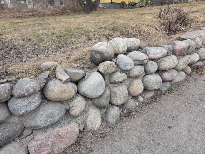 Stenmur av rundade stenar i behov av reparation sträcker sig längs en grusväg, med gräs och buskar i bakgrunden.