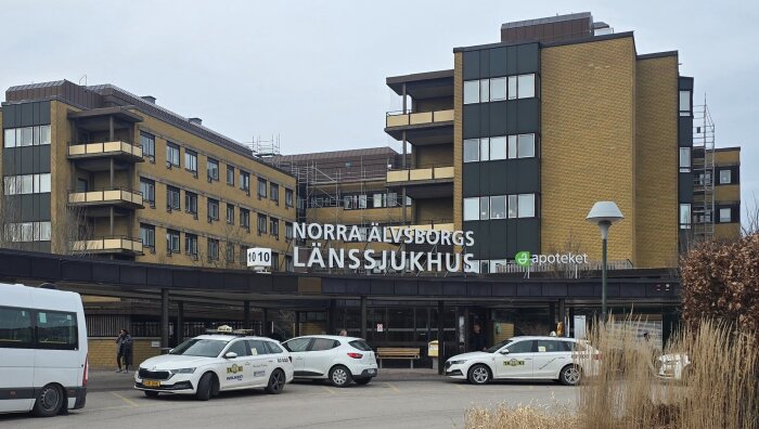 Entrén till Norra Älvsborgs Länssjukhus med taxibilar parkerade framför byggnaden, samt apoteket i bakgrunden.