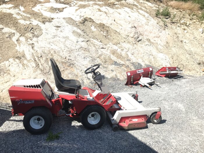 Röd Ventrac redskapsbärare (3 serie) med hydrauliskt tillbehör på grusig mark framför en bergvägg, bredvid några lösa tillbehör.