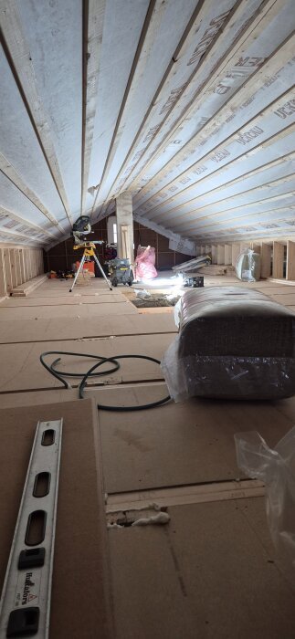Loftutrymme under renovering med synliga innerväggar och takfönster, samt byggmaterial och verktyg i förgrunden.