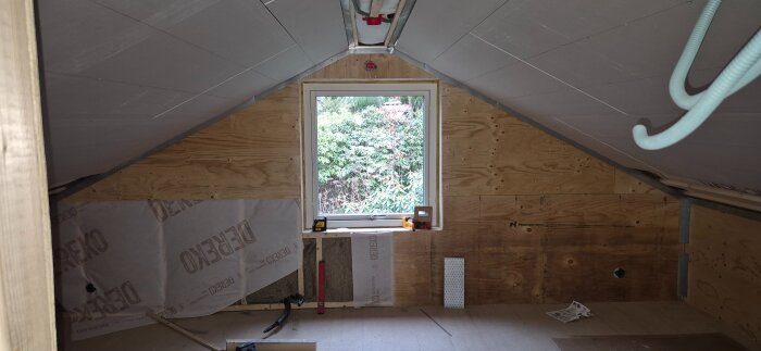 Byggprojekt med innerväggar och ett takfönster under installation, material och verktyg synliga, utsikt mot trädgård genom fönster.