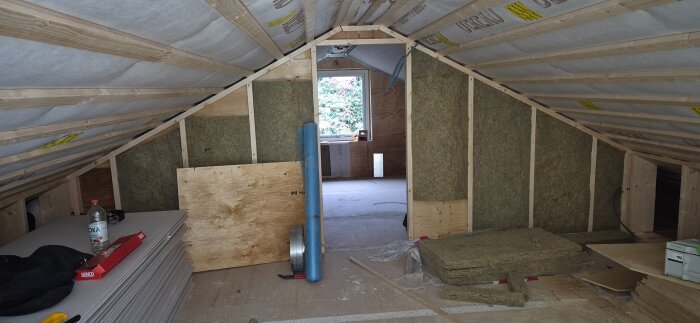 Byggarbetsplats med innerväggar och isolering, takfönster synligt i bakgrunden. Byggmaterial och verktyg på golvet.