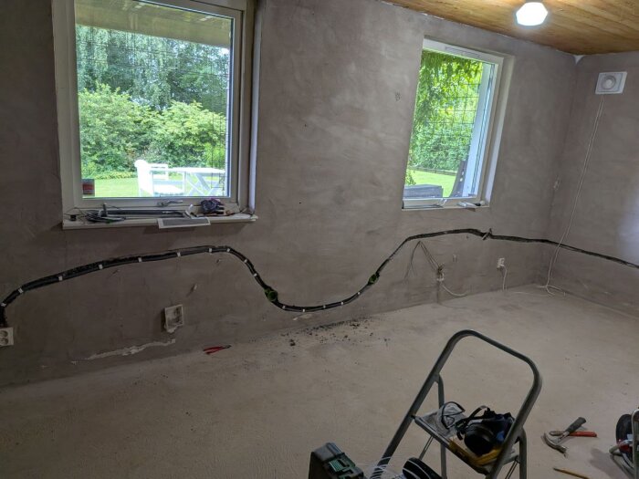 Ett rum under renovering med synliga ledningar i väggarna, två fönster och verktyg på en stege.