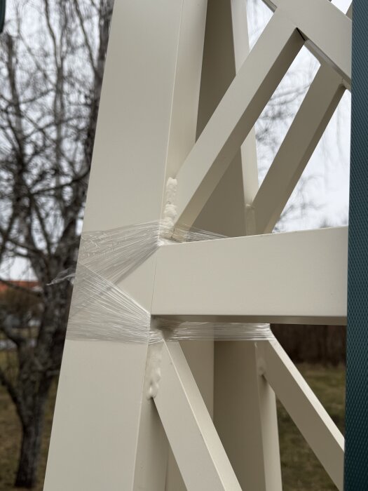 Beige metallstruktur utomhus med plastfolie runt, vid ett träd i bakgrunden.