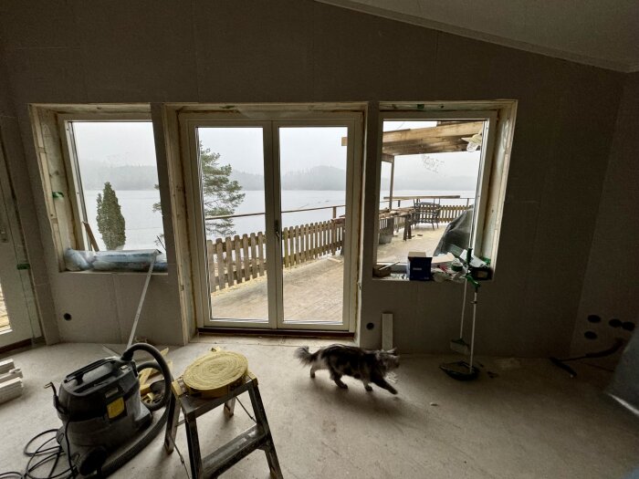 Interiör av ett hus under renovering med utsikt över ett staket och sjö. Ett verktyg och en katt syns på golvet.