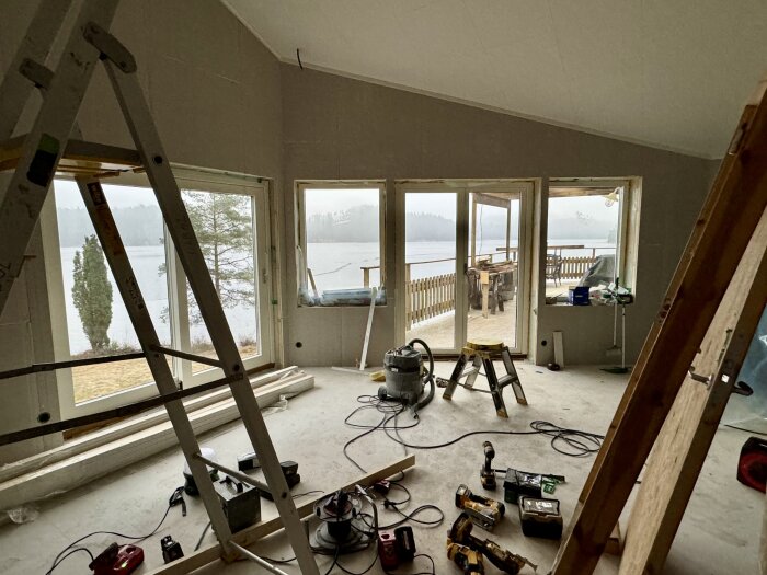 Interiör av byggprojekt med stora fönster mot en sjö, stegar, verktyg och byggmaterial på golvet.