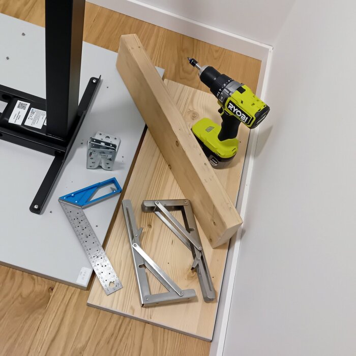 Borrmaskin, träbitar, vinkelhake och beslag på golvet, omgiven av byggmaterial och verktyg.