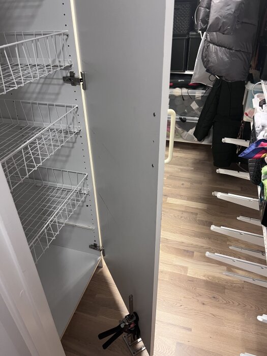 Öppen garderob med vit dörr i klädkammare, med trähylla och hängande kläder. Dörren kapad för att undvika blockering, verktyg synligt.