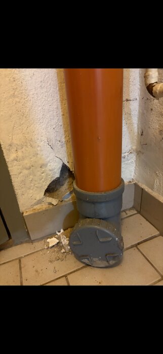 Spricka i murbruket intill ett orange avloppsrör i en källare. Flagnande puts och skräp på golvet vid väggen.