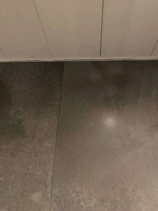 Sprickor i fogen mellan golvplattor och väggpanel i ett badrum.