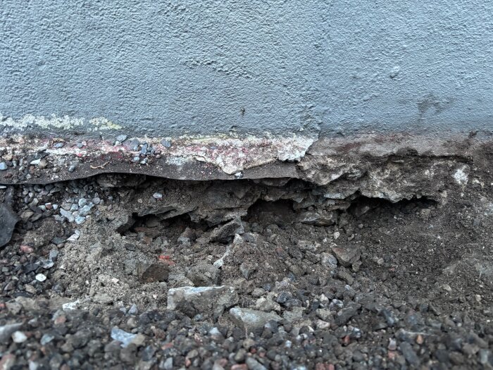 Betonggrund med skador och erosion synlig vid ett hus, omgiven av jord och småsten.