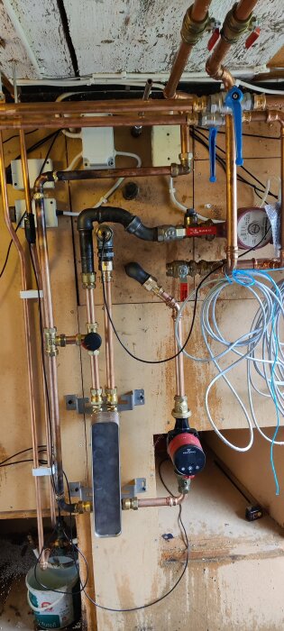 Komplex VVS-installation med kopparrör, ventiler och en cirkulationspump på en vägg med träram, bredvid elektriska kablar.