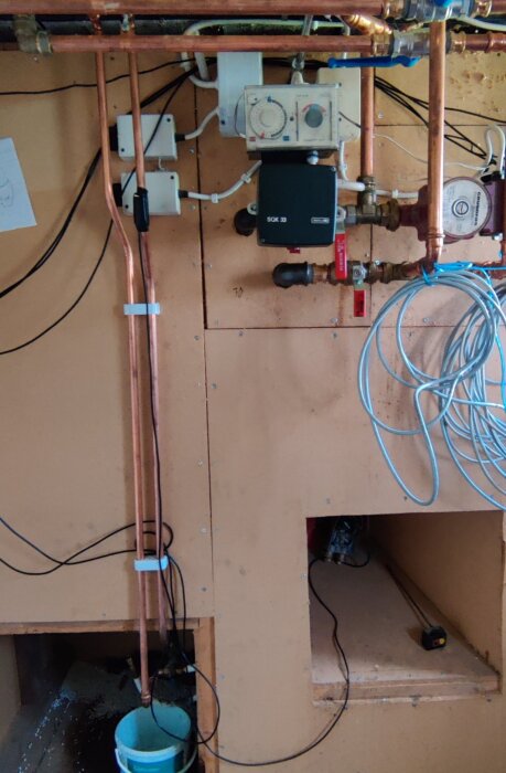 Väggmonterade vattenrör och regulatorer i ett tekniskt utrymme, med kopparrör och elkablar, samt en mätare och ventil installerade.