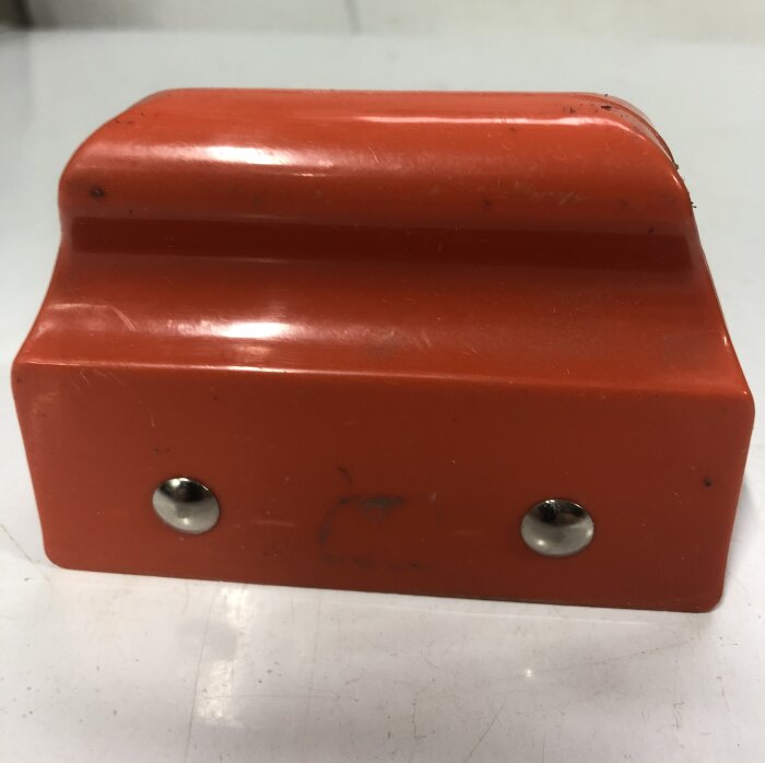 Röd plastenhet med två metallskruvar, oklart ändamål.