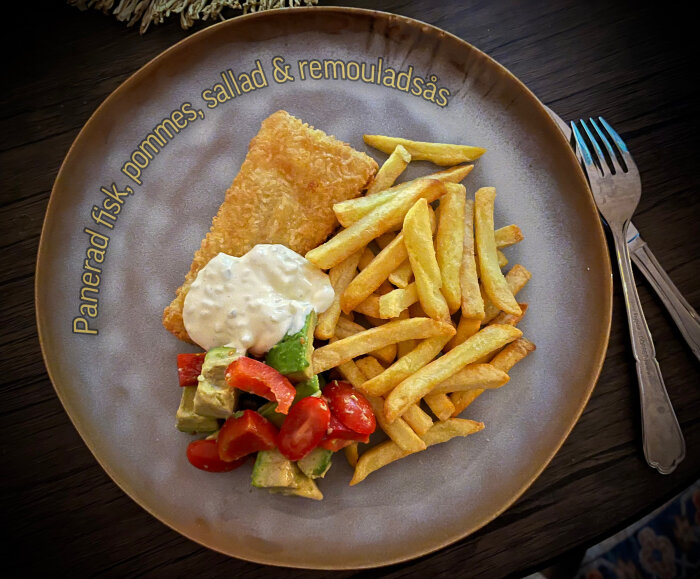 Tallrik med panerad fisk, pommes frites, sallad av avokado och tomat samt remouladsås.