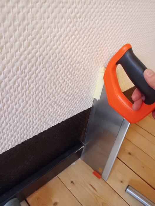 Handsåg med orange handtag kapar list, maskeringstejp skyddar väggen. Golv av träplankor. Förberedelser för garderobsbygge.