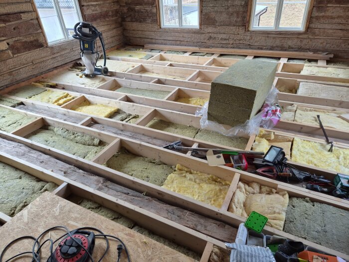 Renoveringsprojekt i gamla huset: Frigjorda bjälkar, isolering, verktyg och dammsugare synliga i pågående golvrenovering.