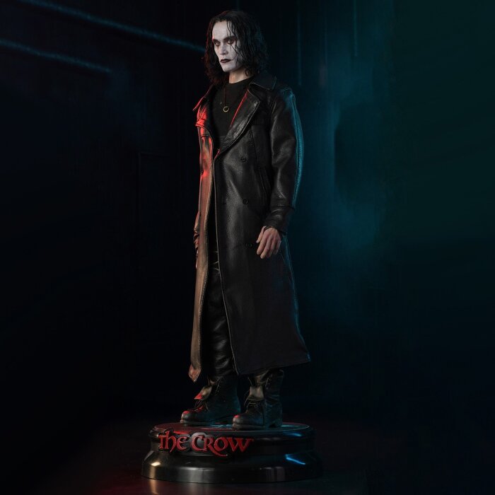 Actionfigur med långt mörkt hår och trenchcoat på ett stativ märkt "The Crow", framhävd mot en mörk bakgrund.