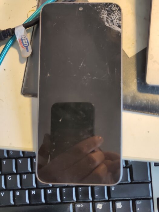 Smartphone med sprucken skärm ovanpå tangentbord, USB-kabel synlig.