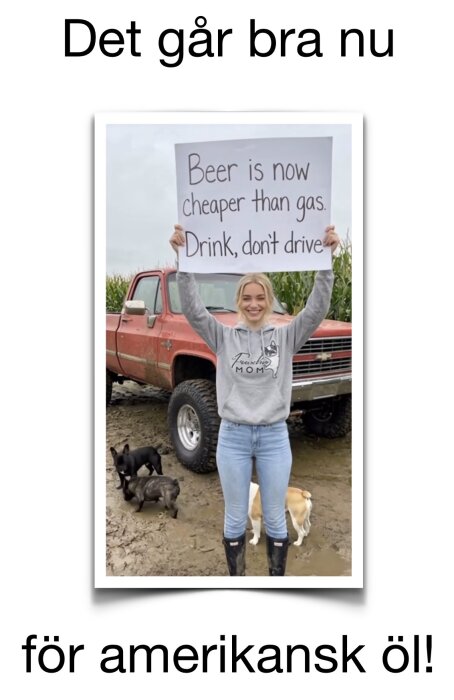 Kvinna håller skylt med texten "Beer is now cheaper than gas. Drink, don't drive" framför en röd pickup, hundar leker i leran.
