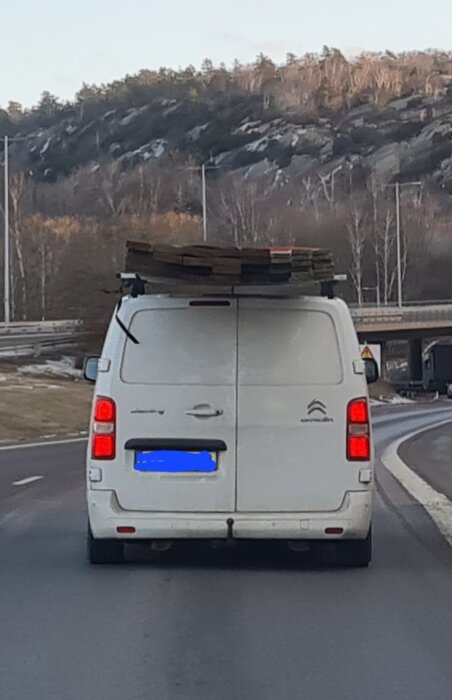 Vit skåpbil med plankor lastade på taket kör på landsväg med skog och berg i bakgrunden.