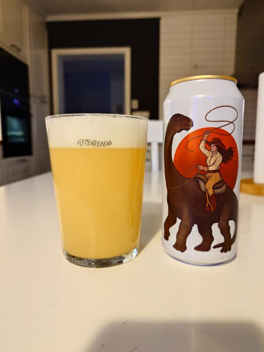 Ett glas öl från Fermenterarna och en ölburk med illustration av en kvinna som rider på en dinosaurie, står på ett vitt bord i ett kök.