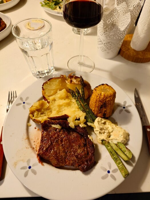 Tallrik med grillad entrecote, bakad potatis, majskolv, sparris och sås. Ett glas rött vin och ett glas vatten står bredvid.