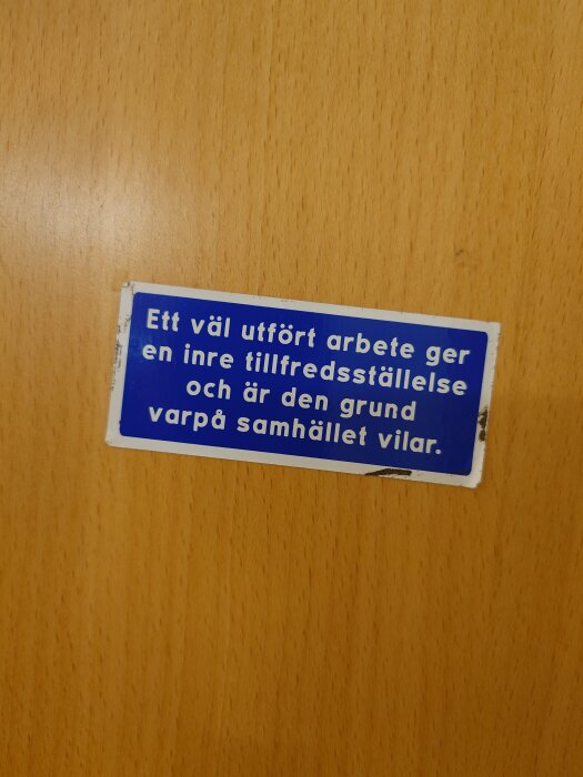 Blå och vit skylt på en träyta med texten: "Ett väl utfört arbete ger en inre tillfredsställelse och är den grund varpå samhället vilar.