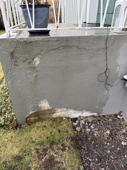 Sliten betongvägg med avflagad färg vid en veranda, omgiven av gräs och grus. En blomkruka står ovanpå väggen.