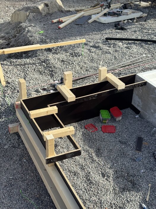 Formar och armeringsjärn på grusig byggarbetsplats, träkonstruktioner förbereder för gjutning av betonggolv. Verktyg och material syns i bakgrunden.