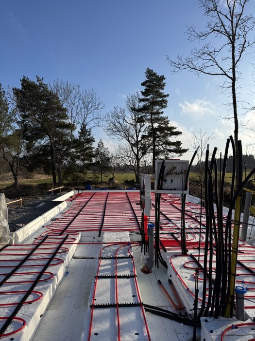 Byggarbetsplats med golvvärmerör installerade på isolering, redo för armering och gjutning av ett synligt betonggolv, med träd och blå himmel i bakgrunden.
