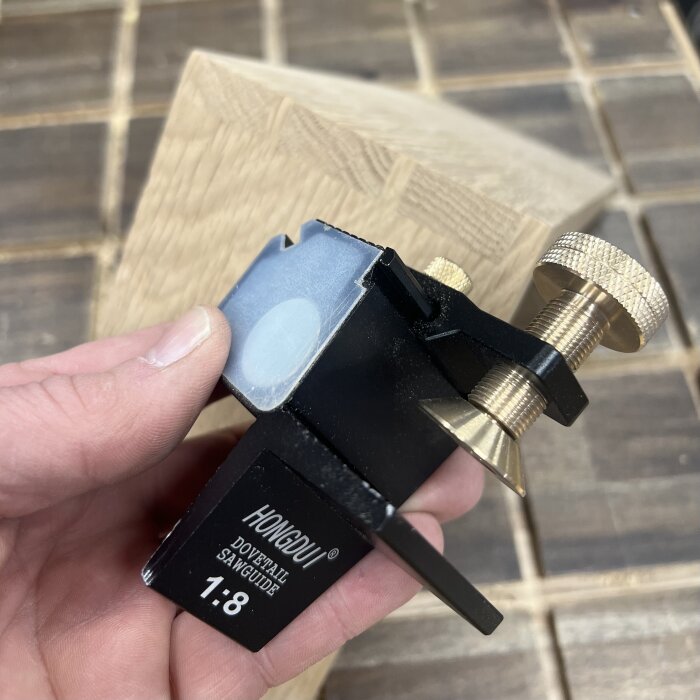 Hand håller en Hongdui dovetail saw guide, en mall för att såga snickarfogar, med en bit trä i bakgrunden på ett verkstadsbord.