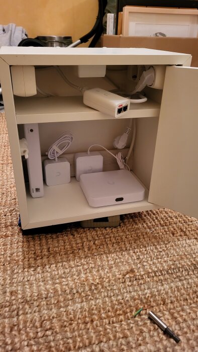 IKEA-låda med Unifi-nätverksutrustning, kablar och transformatorer på hyllor, öppen dörr, står på en matta.