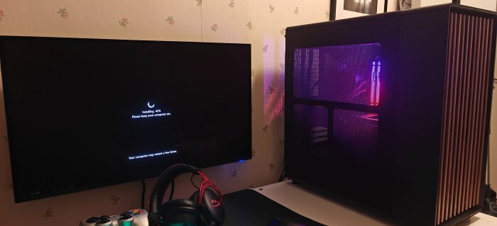 Tv-skärm med installationsmeddelande bredvid en datorlåda med ljuseffekter, headset och spelkontroll på skrivbordet.