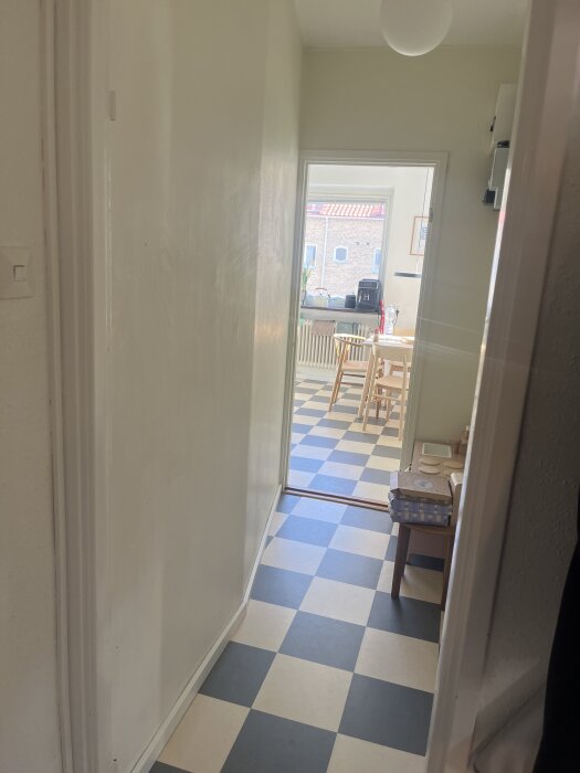 En lång, smal korridor med rutigt golv som leder in i ett kök med bord och stolar synliga i bakgrunden.