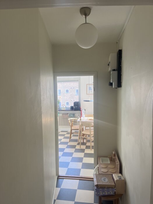 En smal korridor leder in till ett ljust kök med rutigt golv och matbord, där en barnleksaksspis står mot väggen.